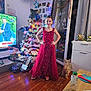 child, girl, red_dress, christmas_tree, christmas_lights, gifts, dog, living_room, television, floor, glass_door, holiday_decor, ornaments, furniture, person, flooring, decorations, smiling, standing, indoors