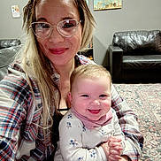 Preszley is registered to the contest to win money with this photo: adult, baby, infant, woman, smiling, glasses, tattoo, plaid_shirt, holding_baby, living_room, couch, area_rug, paintings, portrait, close_up, happy, hands, long_hair, sitting, indoor