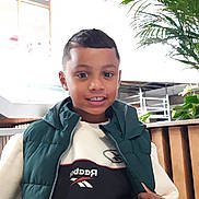 Yvan participe au concours pour gagner de l'argent avec cette photo : child, boy, smiling, indoor, green_puffer_vest, sweatshirt, clothing, face, person, plant, modern_interior, wooden_panel, casual, portrait, young, happy, short_hair, fashion, standing, recreation