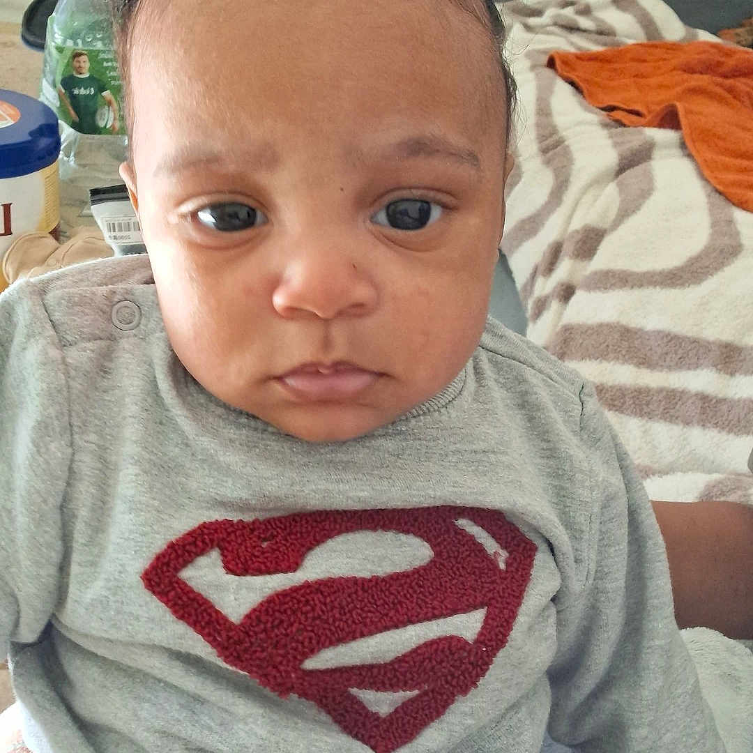 Ynoa participe au concours pour gagner de l'argent avec cette photo : baby, child, superman_logo, gray_sweater, indoor, clothing, blanket, furniture, person, portrait, cute, serious_face, hair_buns, home, toddler, sitting, bottle, laundry, casual, young_child