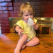 Susan is registered to the contest to win money with this photo: child, toddler, yellow_clothing, curly_hair, table, wood, fireplace, brick_wall, indoors, bottle, cap, sitting, barefoot, cute, face, person, young, chewing, curious, cozy
