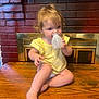 child, toddler, yellow_clothing, curly_hair, table, wood, fireplace, brick_wall, indoors, bottle, cap, sitting, barefoot, cute, face, person, young, chewing, curious, cozy