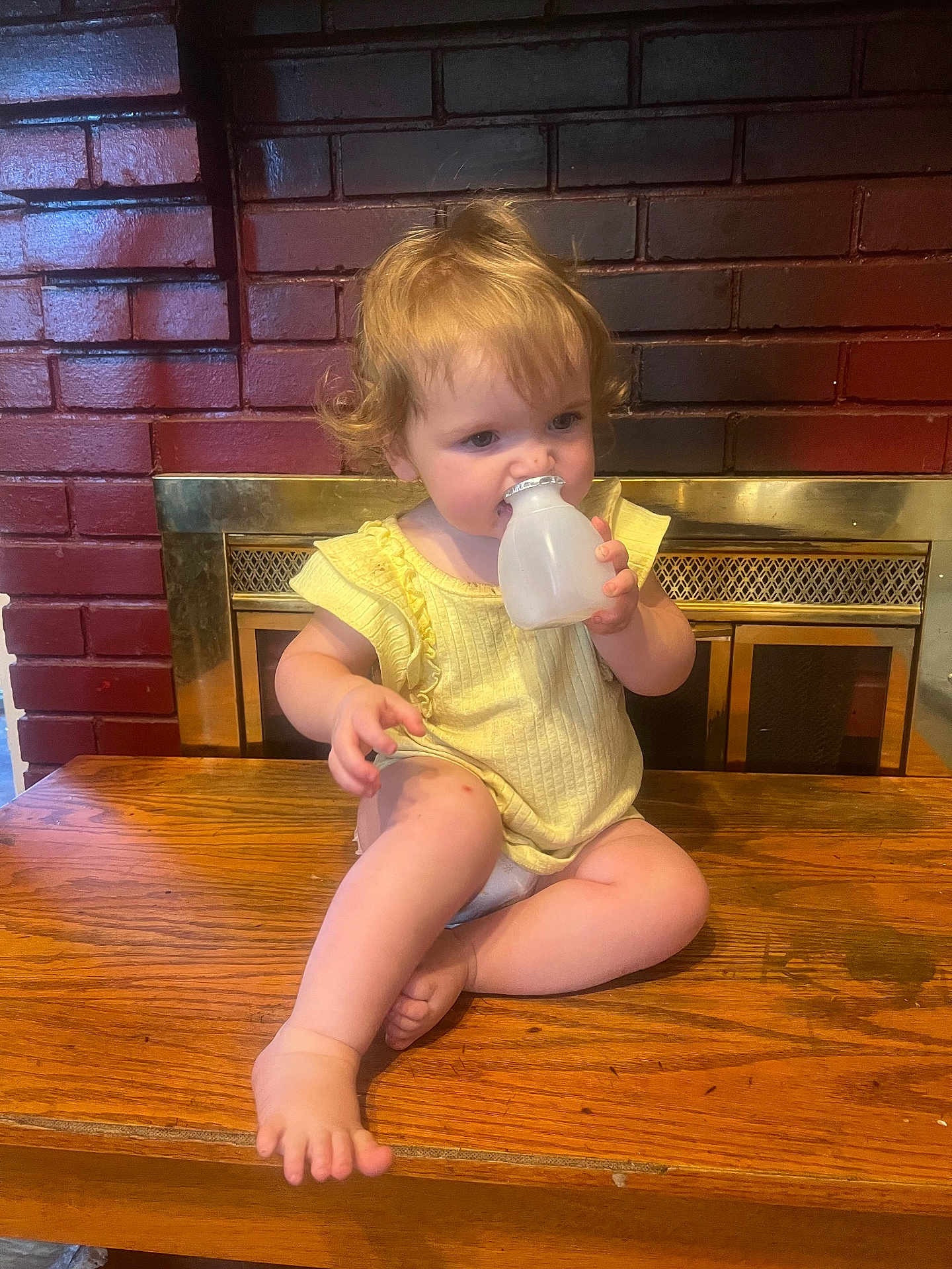 Susan is registered to the contest to win money with this photo: child, toddler, yellow_clothing, curly_hair, table, wood, fireplace, brick_wall, indoors, bottle, cap, sitting, barefoot, cute, face, person, young, chewing, curious, cozy