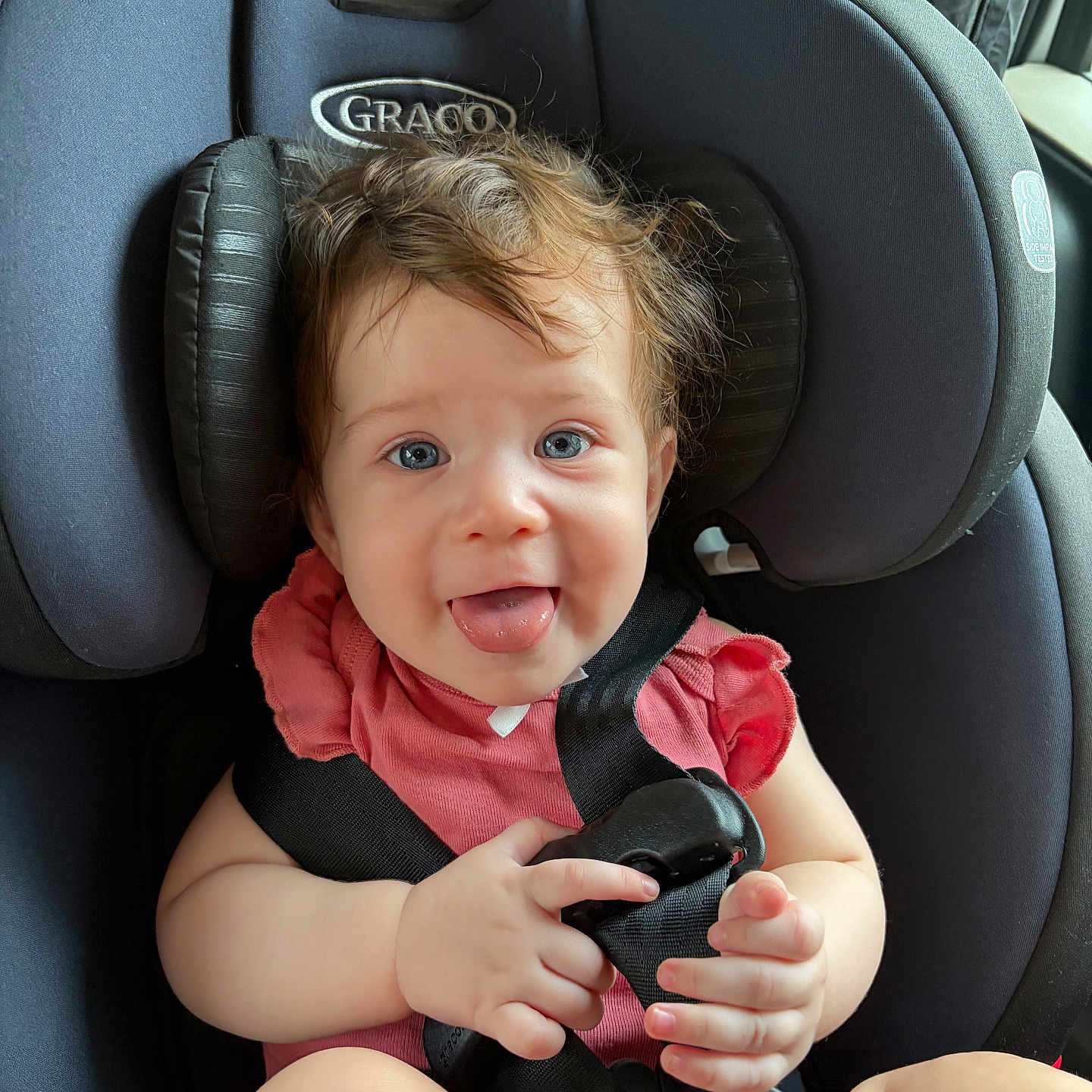 Daysha is registered to the contest to win money with this photo: baby, child, car_seat, seatbelt, blue_eyes, pink_clothing, hair, tongue, hand, infant, smiling, sitting, safety, child_seat, cute, indoors, car, portrait, person, happy