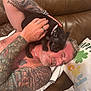 affection, black_dog, casual, chain_necklace, closeup, couch, dog_collar, face, human_hand, indoor, leather, man, pet, pillow, playing, puppy, relaxing, small_dog, tattoo, tattooed_arm