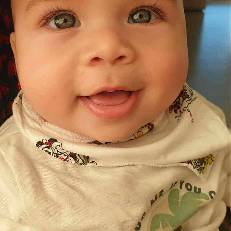 Charaf a rejoint le concours — aidez-le/la à gagner de superbes lots ! adorable, baby, bib, chubby_cheeks, closeup, clothing, eyelashes, eyes, face, happy, indoor, infant, looking_up, person, portrait, shirt, skin, smile, tongue, window