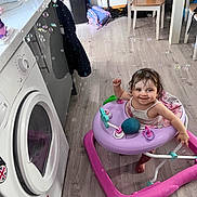 Rosie is registered to the contest to win money with this photo: toddler, walker, bubble, kitchen, floor, appliance, washing_machine, cabinet, chair, table, backpack, smiling, happy, indoor, child, toy, flooring, person, home, play