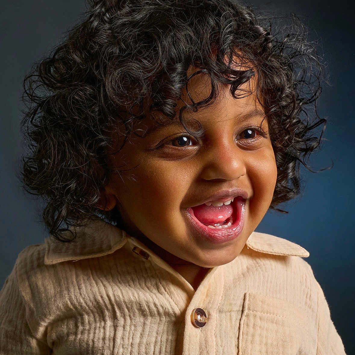 Ayush is registered to the contest to win money with this photo: child, toddler, curly_hair, smiling, laughing, beige_shirt, portrait, happy, cute, face, eyes, mouth, teeth, skin, clothing, person, indoor, studio, dark_background, closeup