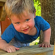 Louis participe au concours pour gagner de l'argent avec cette photo : toddler, child, blue_eyes, blond_hair, outdoor, wooden_structure, tree, playful, smiling, close_up, nature, greenery, daylight, casual_clothing, curious, hands, face, portrait, young_child, summer