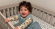 Ezekiel joined the competition — help win amazing prizes! toddler, child, crib, bed, dinosaur_bedding, curly_hair, smiling, happy, indoor, plush_toys, baby_clothes, person, furniture, room, sleeping_area, infant, cute, grip, standing, playful