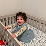 toddler, child, crib, bed, dinosaur_bedding, curly_hair, smiling, happy, indoor, plush_toys, baby_clothes, person, furniture, room, sleeping_area, infant, cute, grip, standing, playful