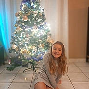 Julia participe au concours pour gagner de l'argent avec cette photo : child, girl, christmas_tree, lights, holiday, decorations, smile, long_hair, floor, socks, indoor, festive, happy, home, seasonal, celebration, tiled_floor, sitting, winter, cozy