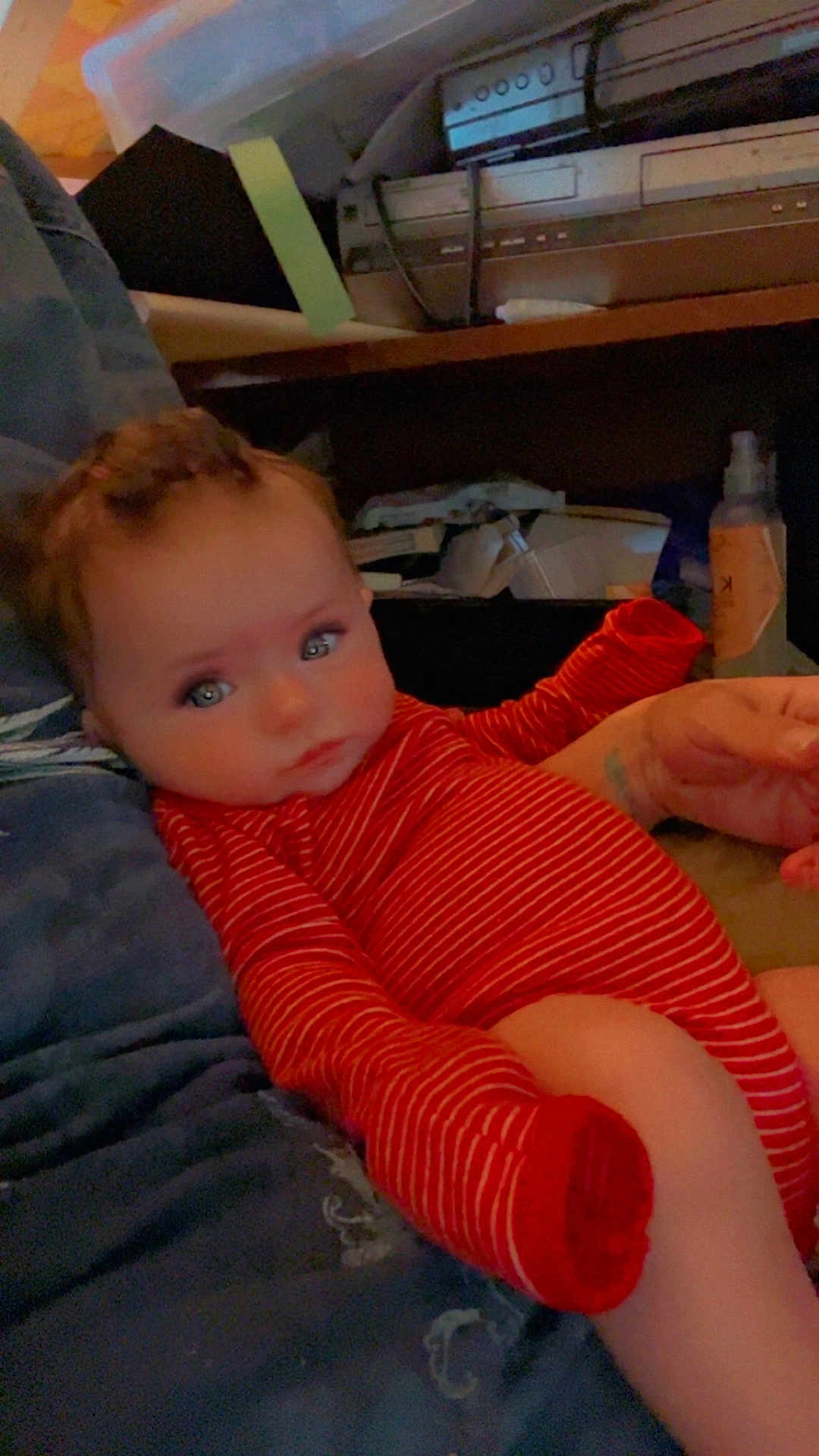 Alexis is registered to the contest to win money with this photo: baby, infant, red_striped_onesie, couch, blue_couch, face, eyes, hair, hand, leg, sitting, indoor, shelf, household_items, clothing, person, portrait, relaxing, child, casual