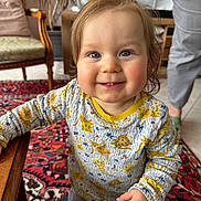 Karol participe au concours pour gagner de l'argent avec cette photo : toddler, child, smile, blue_eyes, long_sleeve_shirt, animal_print, indoor, carpet, furniture, chair, person, hand, footwear, floor_tiles, adult, legs, wooden_table, happy, curly_hair, home