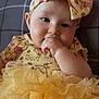 Elliana is registered to the contest to win money with this photo: baby, child, infant, yellow_bow, yellow_dress, ruffles, hand_in_mouth, butterfly_emoji, plaid_background, cute, adorable, headband, smiling, seated, indoors, soft_lighting, face, person, portrait, clothing