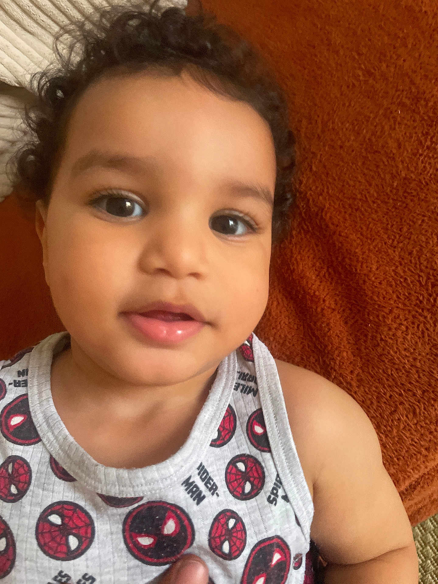 Bayazid a rejoint le concours — aidez-le/la à gagner de superbes lots ! baby, child, toddler, face, curly_hair, tank_top, spiderman, clothing, smile, skin, person, portrait, indoors, closeup, orange_background, cute, young_child, expression, laying_down, casual