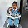 toddler, high_chair, child, sippy_cup, tablet, blue_outfit, sneakers, smile, pointing, indoor, kitchen, floor, light_switch, modern_appliance, person, furniture, clothing, happy, seated, baby