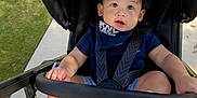 Dylan joined the competition — help win amazing prizes! baby, child, stroller, outdoor, sidewalk, grass, black_stroller, blue_shirt, shorts, barefoot, baby_bib, curious_expression, sunlight, seatbelt, infant, person, young_child, casual_clothing, foot, hand