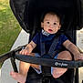 baby, child, stroller, outdoor, sidewalk, grass, black_stroller, blue_shirt, shorts, barefoot, baby_bib, curious_expression, sunlight, seatbelt, infant, person, young_child, casual_clothing, foot, hand