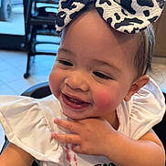 Isabella is registered to the contest to win money with this photo: toddler, child, smiling, headband, bow, cow_print, white_clothing, indoor, high_chair, cute, happy, portrait, baby, hand_near_face, messy, casual, floor_tiles, background, furniture, person