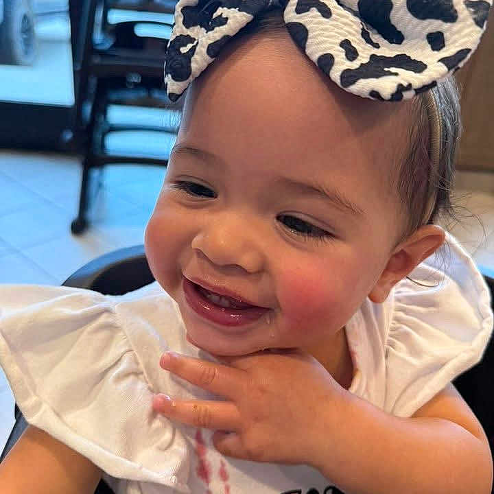 Isabella is registered to the contest to win money with this photo: baby, background, bow, casual, child, cow_print, cute, floor_tiles, furniture, hand_near_face, happy, headband, high_chair, indoor, messy, person, portrait, smiling, toddler, white_clothing