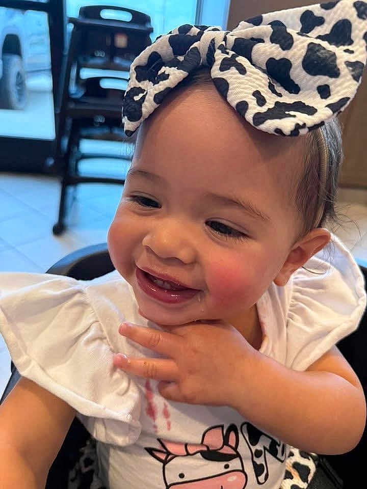 Isabella is registered to the contest to win money with this photo: toddler, child, smiling, headband, bow, cow_print, white_clothing, indoor, high_chair, cute, happy, portrait, baby, hand_near_face, messy, casual, floor_tiles, background, furniture, person