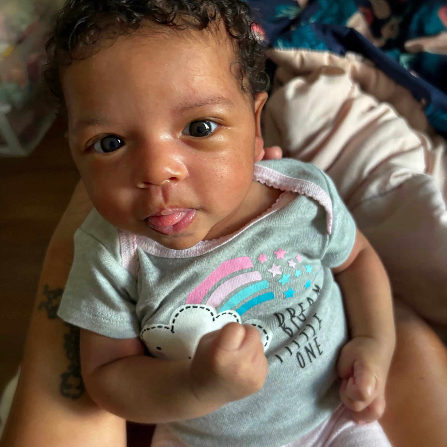 Jayla joined the competition — help win amazing prizes! baby, infant, child, curly_hair, tongue_out, onesie, rainbow, stars, fist, close_up, portrait, cute, playful, person, skin, expression, hand, indoor, holding, cozy