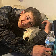 Mickey joined the competition — help win amazing prizes! person, dog, smiling, cuddling, bed, pillow, water_bottle, indoor, casual_clothing, young_man, white_dog, face, hand, sleeve, plaid_shirt, wall, shelf, cozy, relaxed, affection