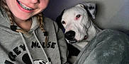 Mickey is registered to the contest to win money with this photo: person, dog, smile, braids, hoodie, mickey_mouse, blanket, indoors, portrait, closeup, cute, pet, friendship, young_adult, casual, relaxed, brown_eyes, nose_ring, black_and_white_dog, cozy