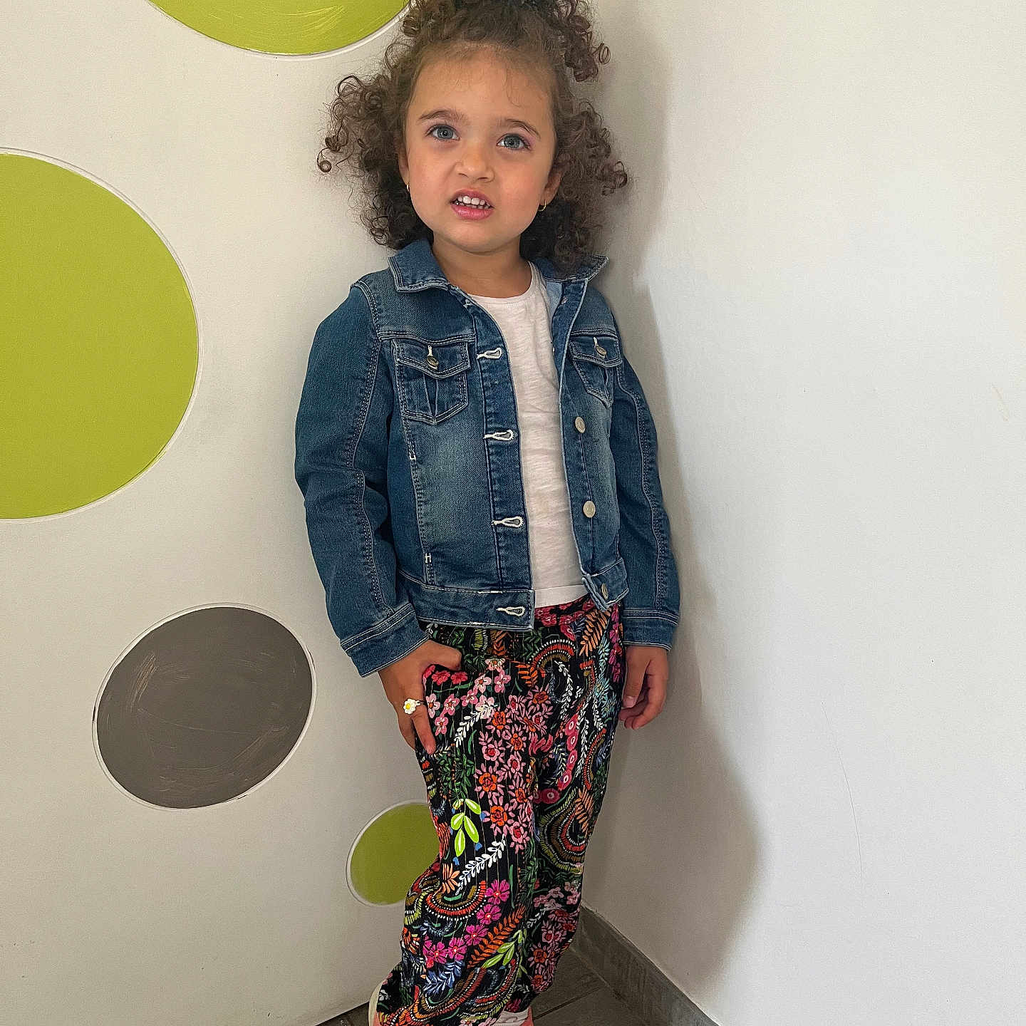 Louna participe au concours pour gagner de l'argent avec cette photo : casual_clothing, child, colorful, curly_hair, cute, decor, denim_jacket, fashion, floor, floral_pants, girl, happy, indoor, person, pink_shoes, portrait, smile, standing, wall, young