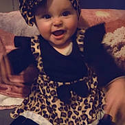 Helyana participe au concours pour gagner de l'argent avec cette photo : baby, infant, smiling, headband, bow, leopard_print, dress, black_top, sitting, couch, blanket, indoors, portrait, happy, big_eyes, chubby_cheeks, hands, legs, footwear, adorable
