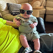 Kayson is registered to the contest to win money with this photo: baby, sunglasses, baby_clothes, adult_arm, couch, dog, living_room, pillow, footwear, person, casual, indoor, relaxing, leather_furniture, child, cute, plush, seated, shoes, pet