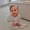 child, toddler, smiling, sitting, indoor, carpet, knitted_sweater, tights, hair_clip, beige, couch, plant, home_interior, happy, cute, portrait, baby, young_child, floor, cozy