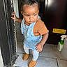 Sernity is registered to the contest to win money with this photo: toddler, child, indoor, doorway, curly_hair, overalls, brown_shirt, white_tights, boots, tile_floor, hand_on_doorframe, exploring, serious_expression, hair_pony_tail, young_child, floor, household, kitchen, person, footwear