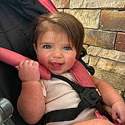 Harlee is registered to the contest to win money with this photo: baby, child, blue_eyes, smiling, stroller, pink_harness, hand, face, skin, cheeks, clothing, seatbelt, safety, person, outdoor, stone_wall, happy, cute, infant, portrait