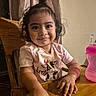 Elizabeth is registered to the contest to win money with this photo: toddler, child, smile, happy, face, curly_hair, pony_tail, pink_shirt, cartoon_cow, wooden_table, crumbs, sippy_cup, pink_cup, indoor, curtain, chair, hand, table, food, casual