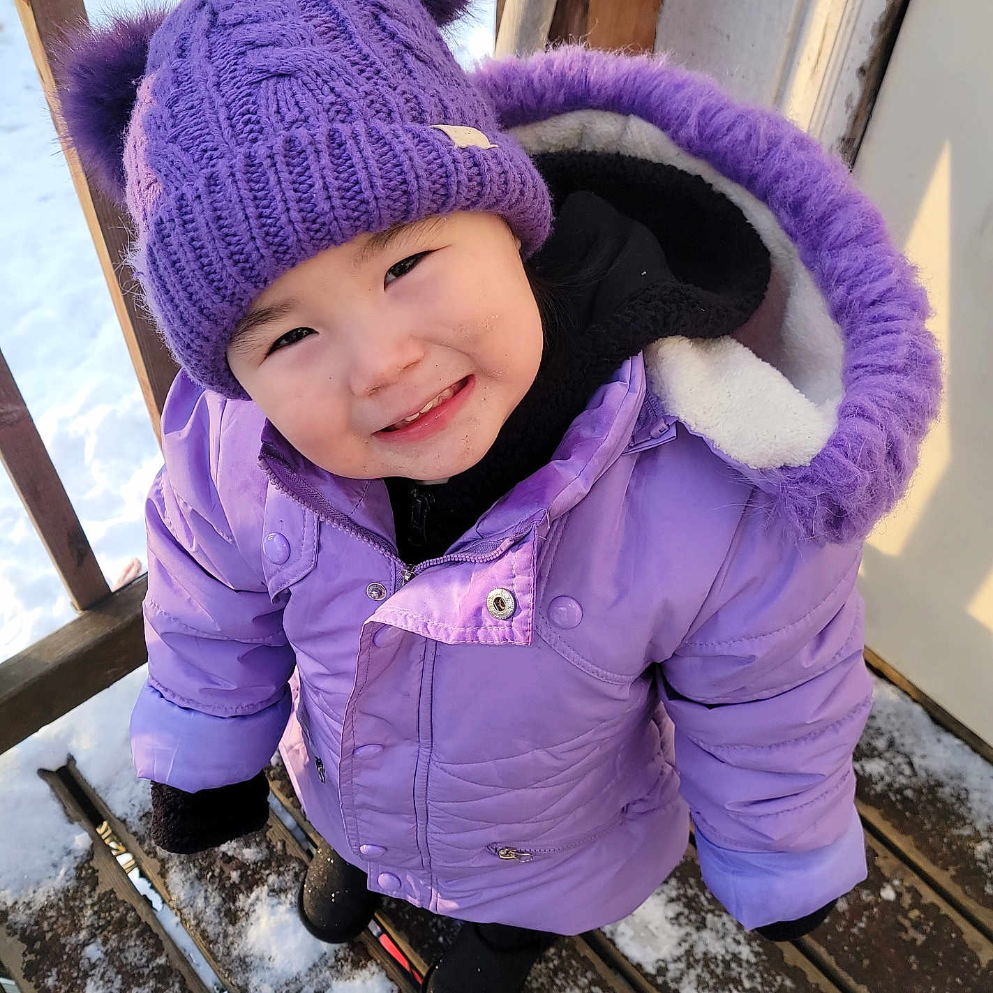 Freya is registered to the contest to win money with this photo: child, toddler, smiling, purple_hat, winter_jacket, snow, outdoor, wooden_deck, cold_weather, skis, gloves, cute, winter_clothing, happy, face, headwear, footwear, playful, person, seasonal