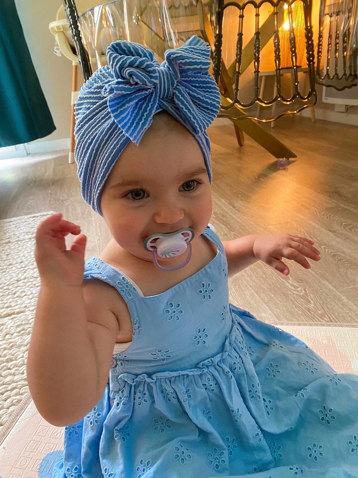 Arya participe au concours pour gagner de l'argent avec cette photo : toddler, child, baby, blue_dress, bow_turban, pacifier, indoor, wooden_floor, carpet, arm, face, headwear, sitting, cute, person, furniture, chair, lighting, home, smile