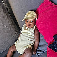 Ahmed participe au concours pour gagner de l'argent avec cette photo : baby, child, cap, hat, couch, cushion, red_cushion, gray_cushion, infant, person, relaxing, indoors, clothing, sleeveless_outfit, barefoot, skin, face, looking, seated, comfortable
