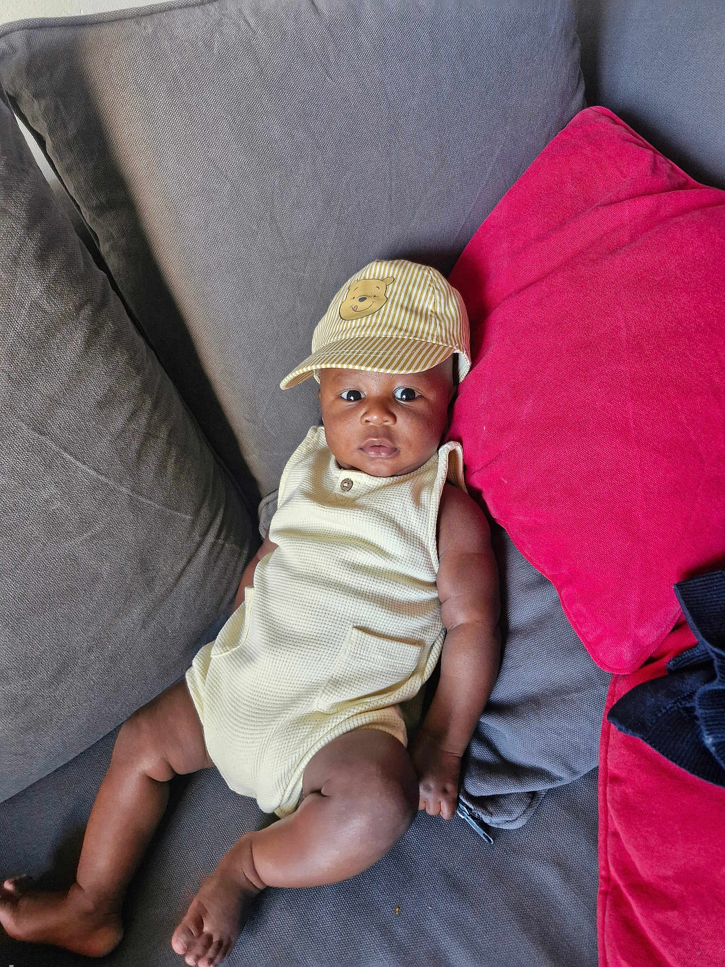 Ahmed participe au concours pour gagner de l'argent avec cette photo : baby, child, cap, hat, couch, cushion, red_cushion, gray_cushion, infant, person, relaxing, indoors, clothing, sleeveless_outfit, barefoot, skin, face, looking, seated, comfortable