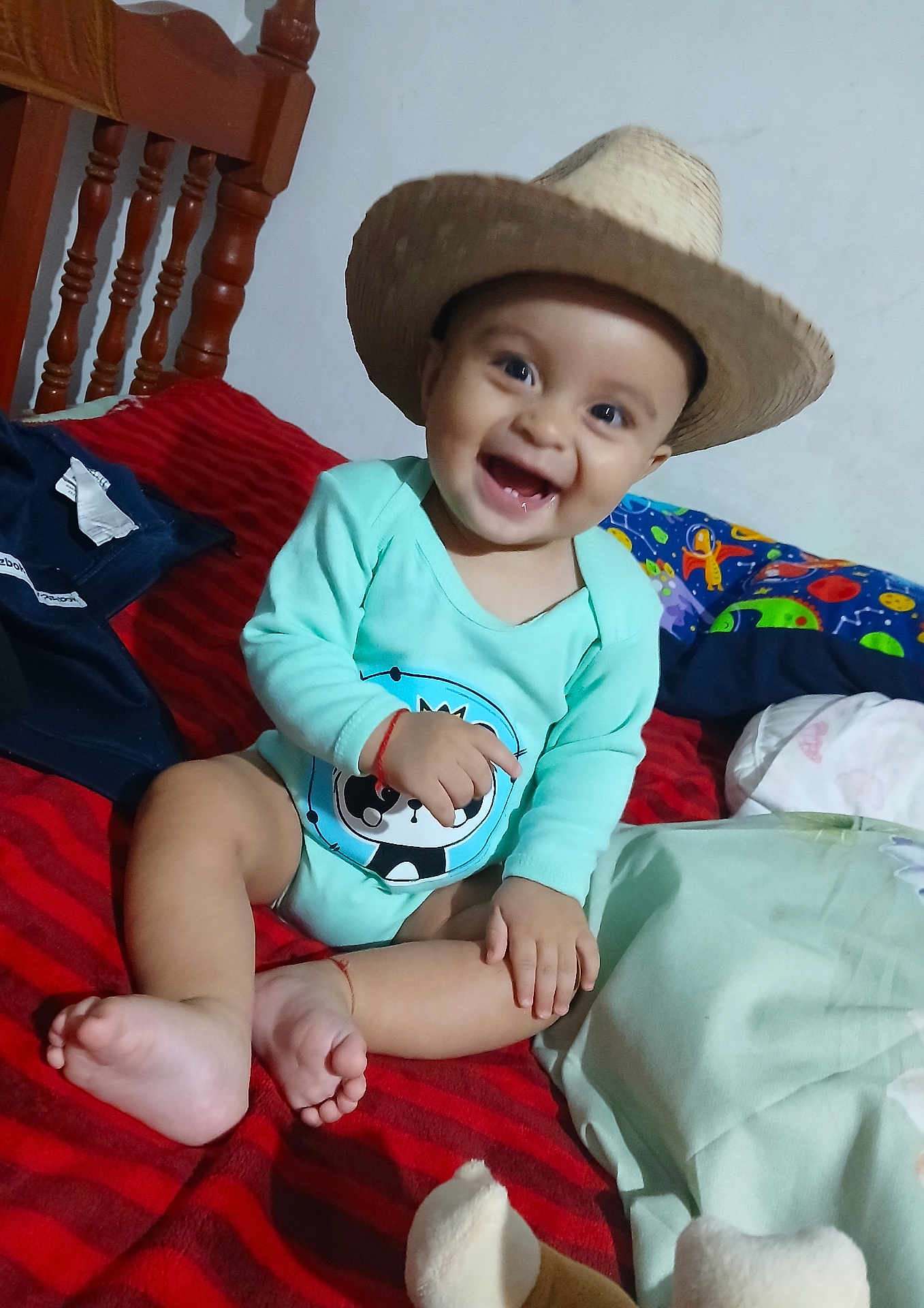 Adan joined the competition — help win amazing prizes! baby, child, smiling, hat, onesie, panda_print, bed, red_stripes, wooden_chair, indoor, happy, cute, infant, sitting, footwear_absent, home, toy, colorful_bedding, playful, portrait