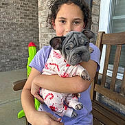 Shelby is registered to the contest to win money with this photo: bench, brick_wall, casual, child, clothing, cozy, curly_hair, cute, dog, french_bulldog, happy, holding, outdoor, pajamas, pet, porch, puppy, smiling, toy, young