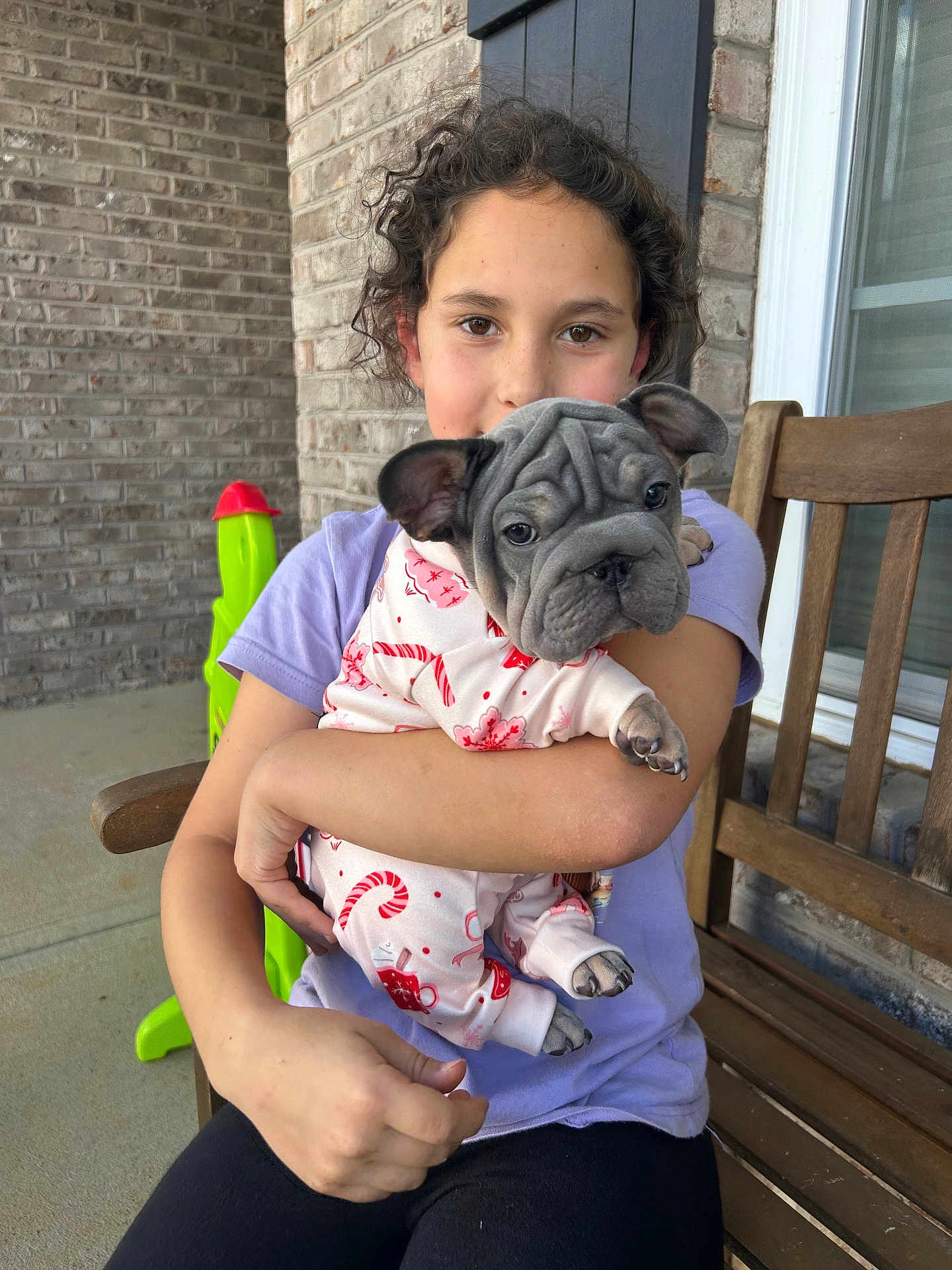 Shelby is registered to the contest to win money with this photo: bench, brick_wall, casual, child, clothing, cozy, curly_hair, cute, dog, french_bulldog, happy, holding, outdoor, pajamas, pet, porch, puppy, smiling, toy, young