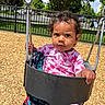 toddler, child, swing, playground, outdoor, curly_hair, pink_clothing, heart_pattern, blanket, wood_chips, fence, trees, sunny_day, park, hand, face, expression, seat, metal_chain, nature