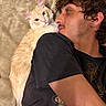 cat, man, human, pet, cream_cat, fluffy, long_hair, curly_hair, portrait, selfie, indoors, bed, lying_down, affection, kissing, animal, feline, black_shirt, closeup, cozy
