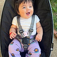 Leilany joined the competition — help win amazing prizes! baby, child, pigtails, hair_ties, baby_seat, black_cushion, white_shirt, purple_pants, sunflower_pattern, socks, outdoor, grass, concrete, happy, smiling, seated, infant, baby_accessory, playful, cute