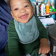 Ezekiel joined the competition — help win amazing prizes! baby, child, smiling, green_clothing, bib, indoor, person, face, happy, sitting, table, bottle, paper, mirror, hair, skin, room, furniture, toy, clutter