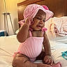 baby, child, infant, smiling, pink_clothing, striped_clothing, hat, bonnet, sitting, bed, blanket, headboard, furniture, indoor, cute, happy, skin, hand, foot, person