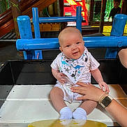 Rohan is registered to the contest to win money with this photo: baby, child, dinosaur_bib, bench, hand, playground, indoor_play_area, colorful, wooden_bench, smiling, socks, person, toy_area, netting, background_child, play_structure, casual_clothing, watching, happy, support