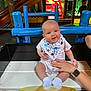 baby, child, dinosaur_bib, bench, hand, playground, indoor_play_area, colorful, wooden_bench, smiling, socks, person, toy_area, netting, background_child, play_structure, casual_clothing, watching, happy, support